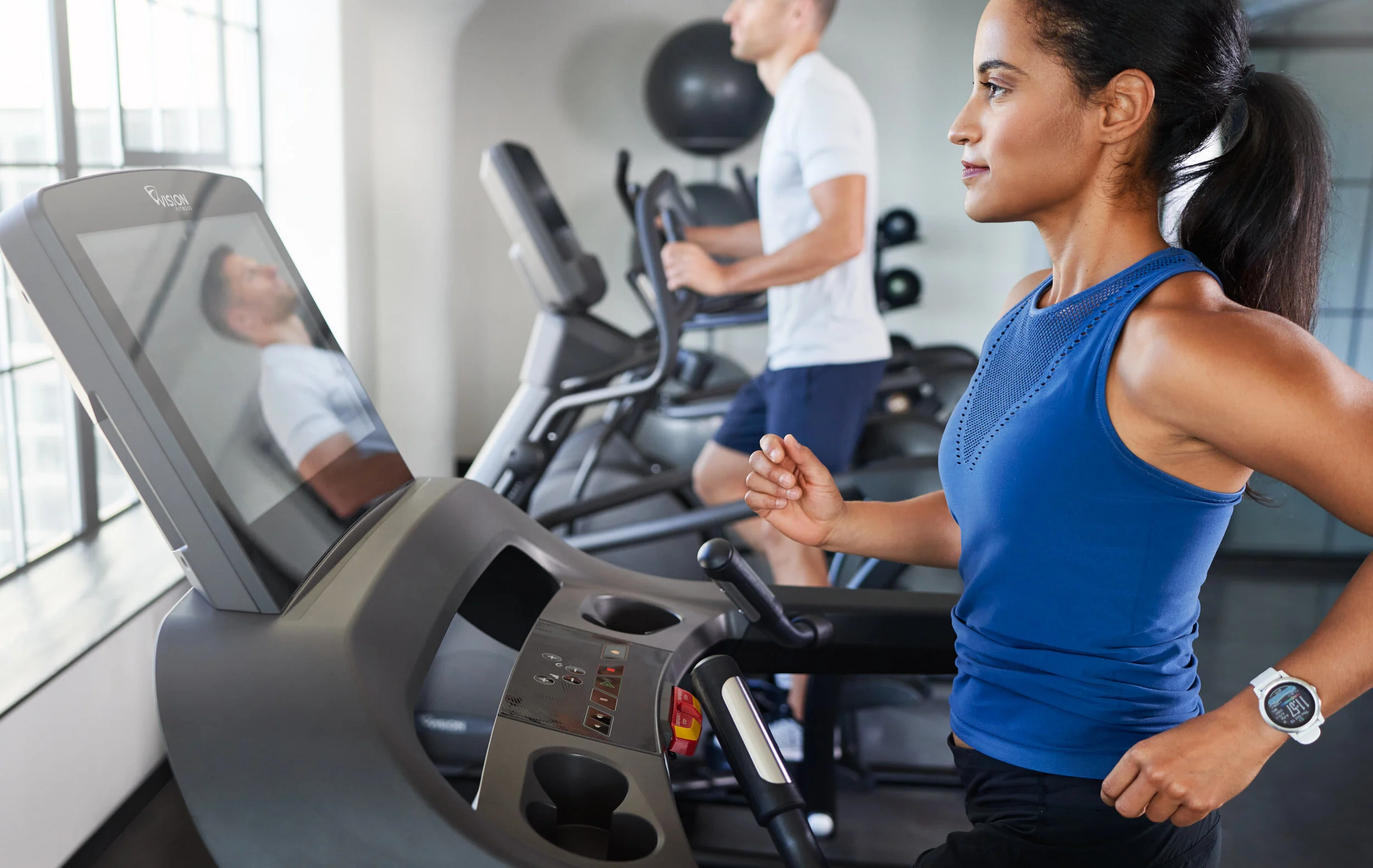A woman running on a treadmill in a gym with a man on a different treadmill in the background.