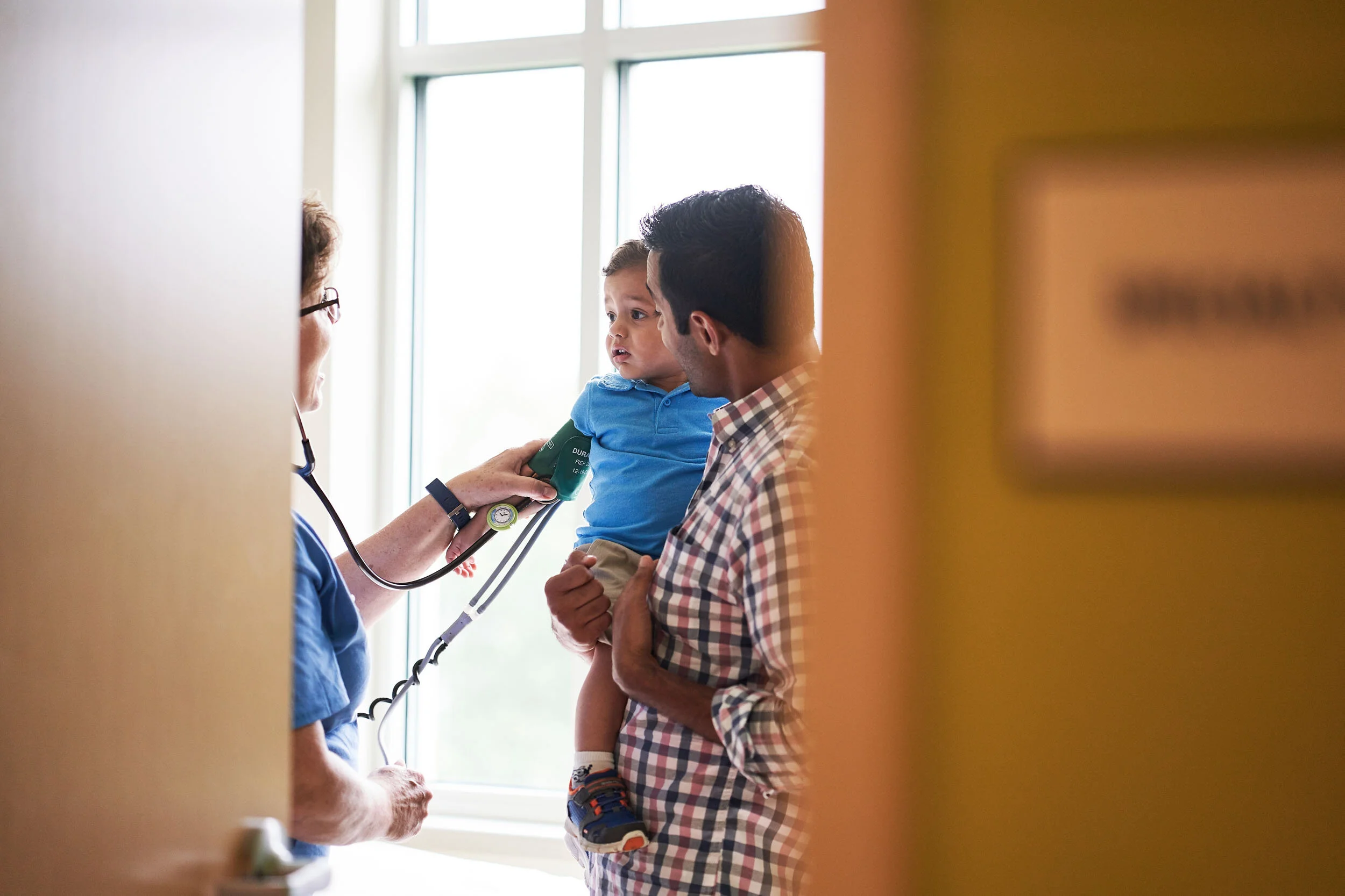 A young boy is having his blood pressure checked by a nurse in a hospital, while his father holds him.