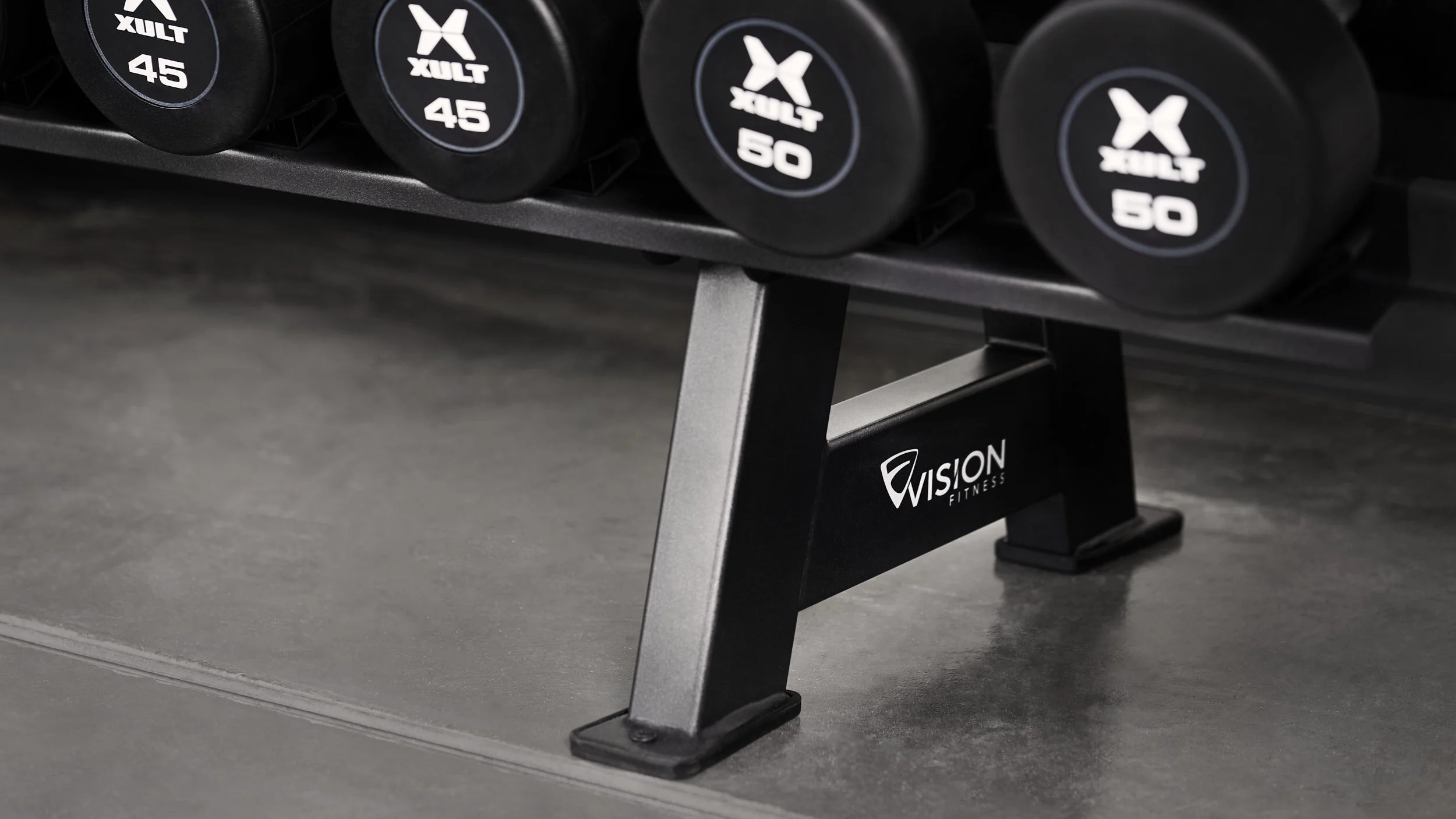 A close-up of a black gym dumbbell rack with weights labeled Xult 45, 50, and 55 pounds, supported by a black and silver Vision Fitness bench leg on a smooth gray gym floor.