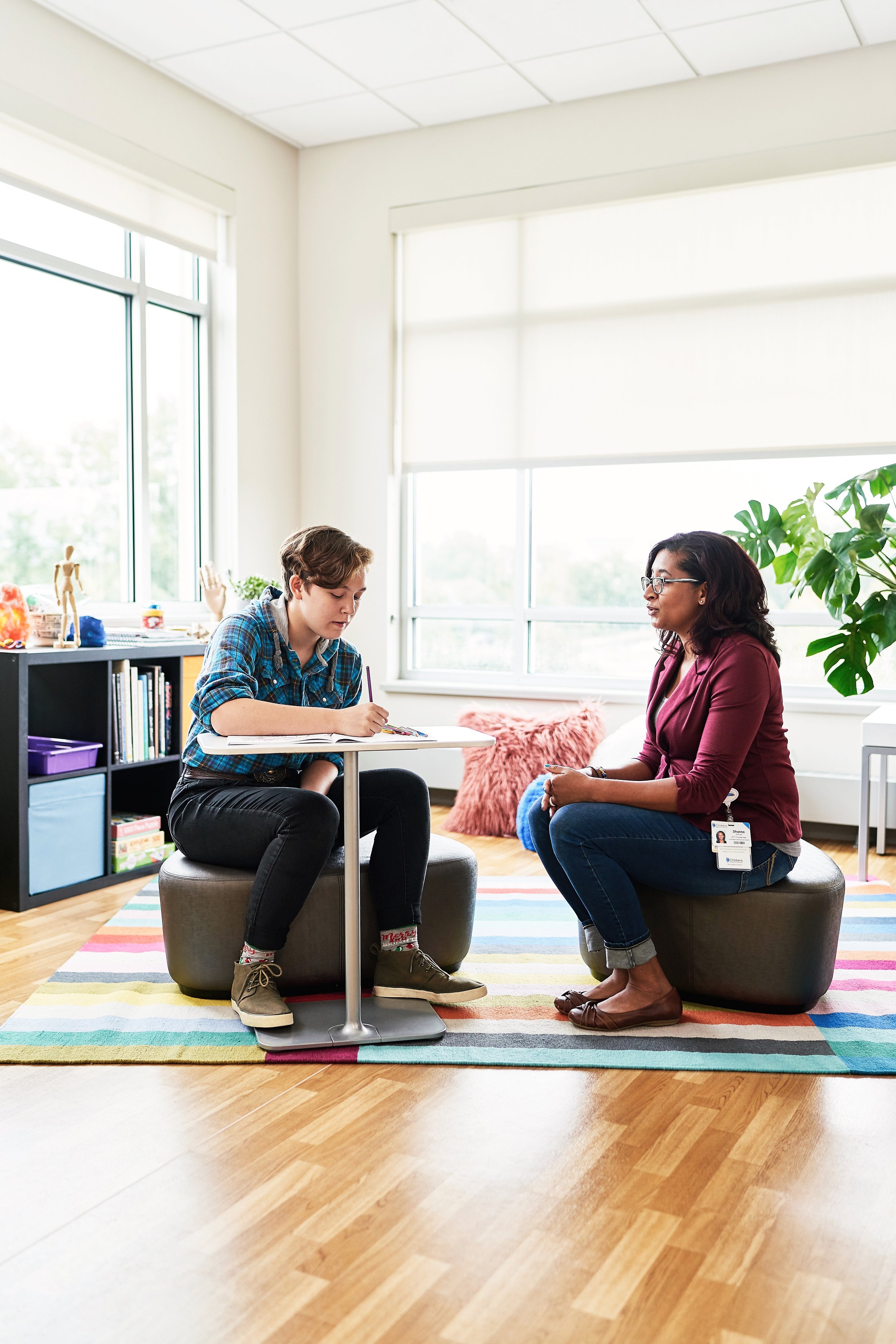 A teenager works in a notebook while sitting across from a mental health therapist in a bright, colorful counseling room at Children's Wisconsin