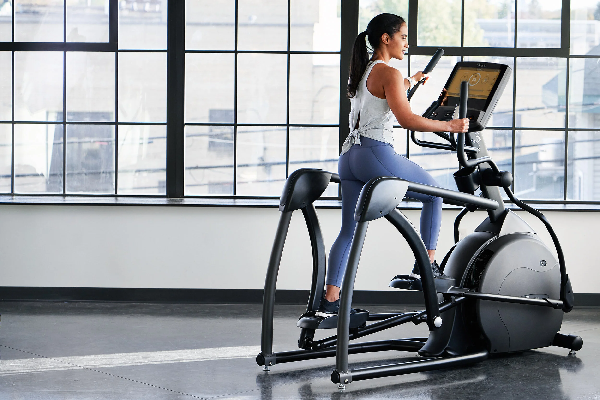 Woman using elliptical machine in a gym with large windows behind.