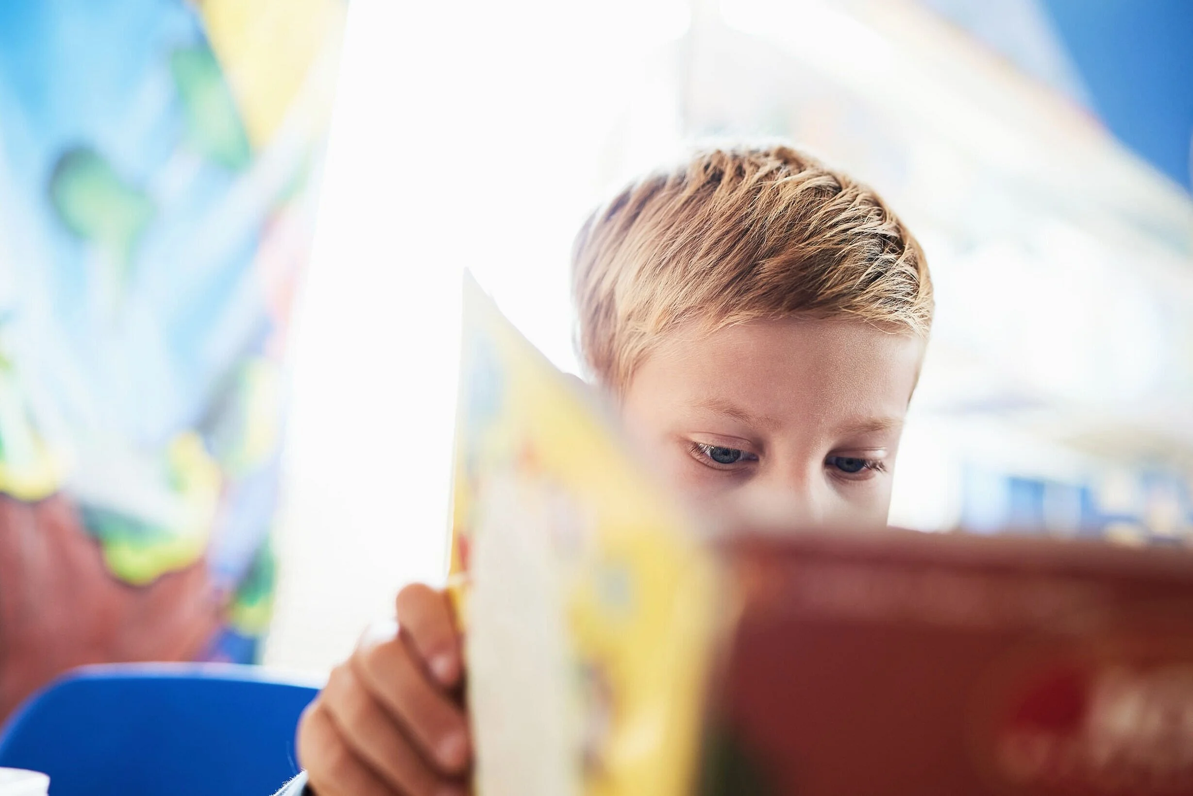 A young boy with blonde hair reading a colorful book, focused on the pages.
