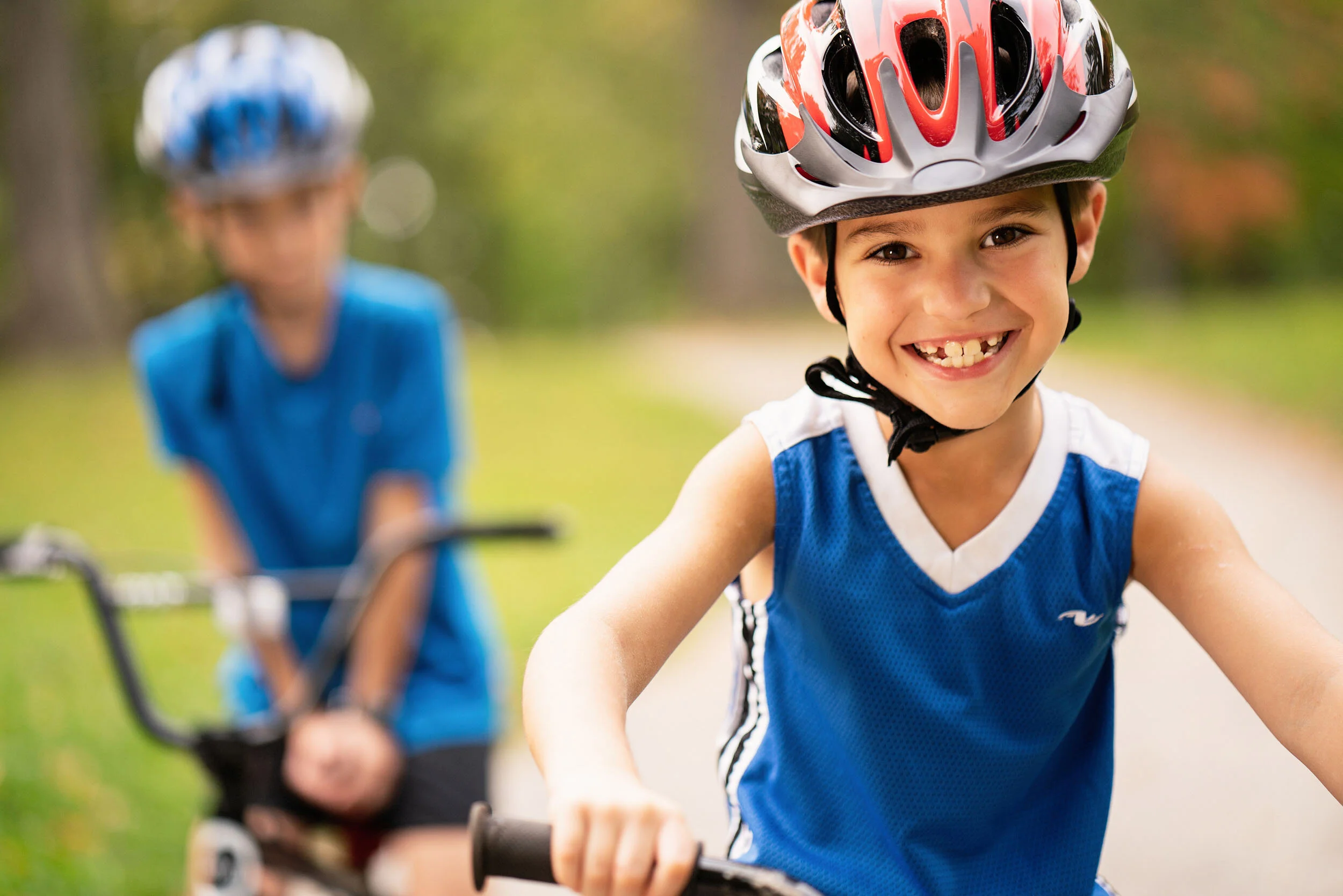 Children riding bikes outdoors, a smiling boy in the foreground wearing a red and gray helmet and a blue sleeveless shirt, with a girl in a blue shirt and a helmet slightly blurred in the background.