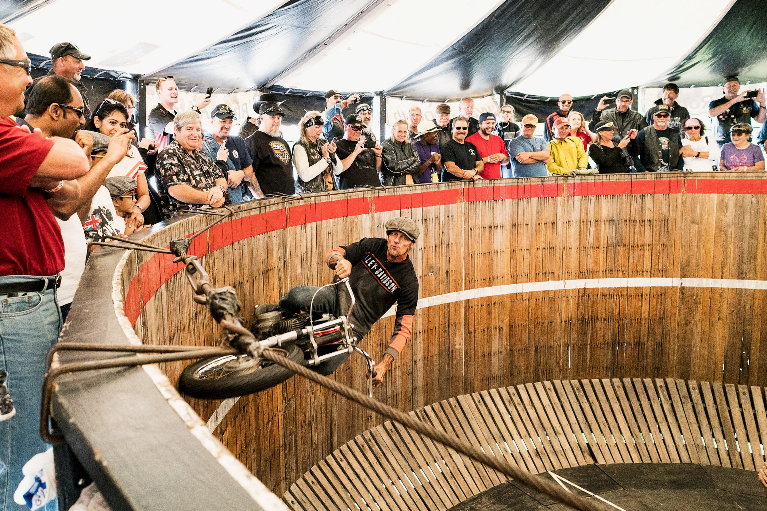 A motorcycle rider performing a stunt inside a wooden, circular arena while a large crowd watches from behind a guardrail.