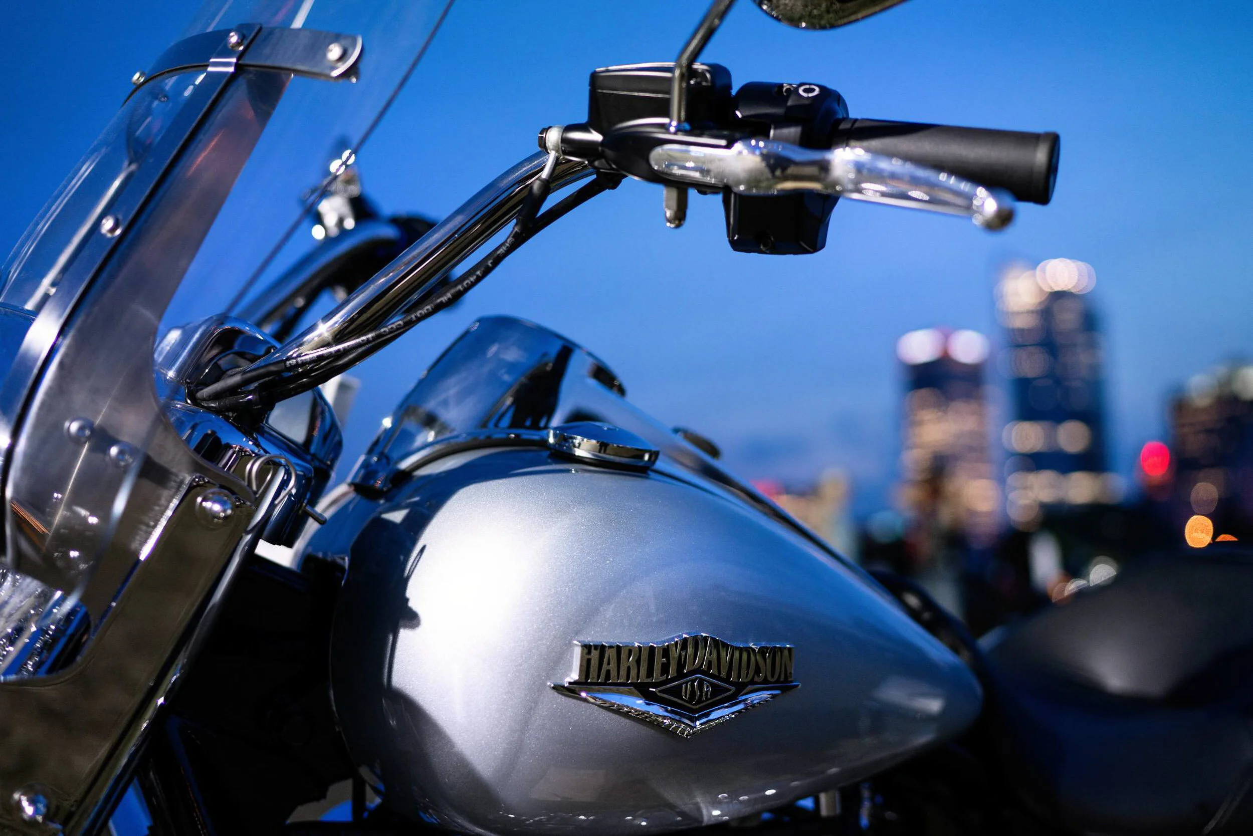 Close-up of a silver Harley Davidson motorcycle with city skyline in the background during dusk.