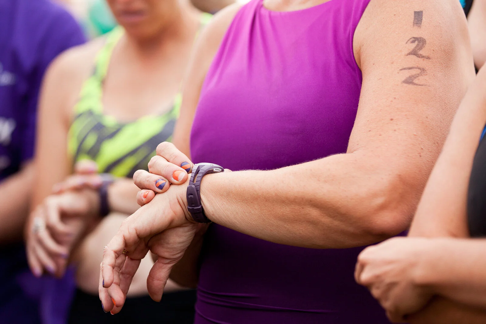 Close-up of a woman wearing a purple sleeveless athletic top, checking her watch with her right hand, with tattoos on her arm. Other women in athletic clothing are in the background.
