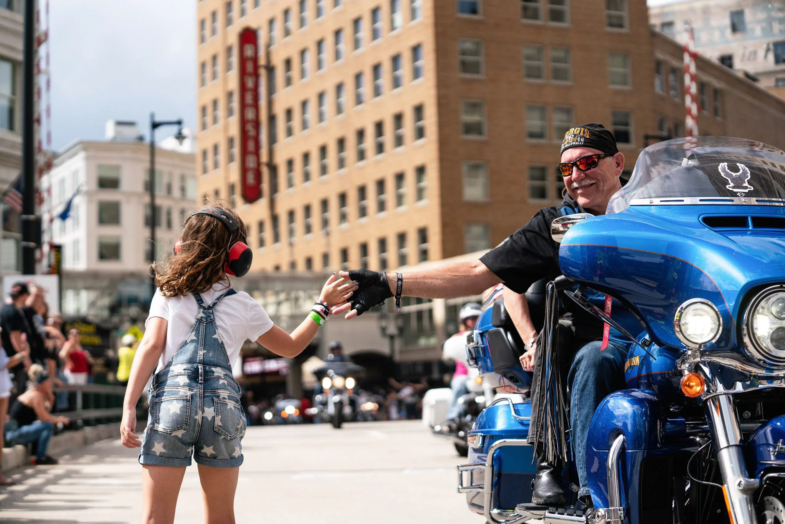 A young girl wearing headphones and denim overalls shaking hands with a smiling man on a blue motorcycle in an urban city street.