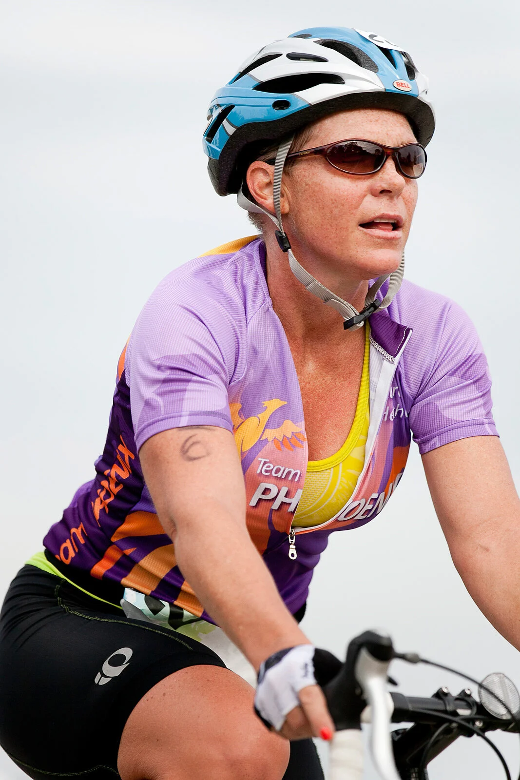 A woman cycling outdoors wearing a purple and yellow cycling jersey, black shorts, a blue and white helmet, and sunglasses, with a focused expression.