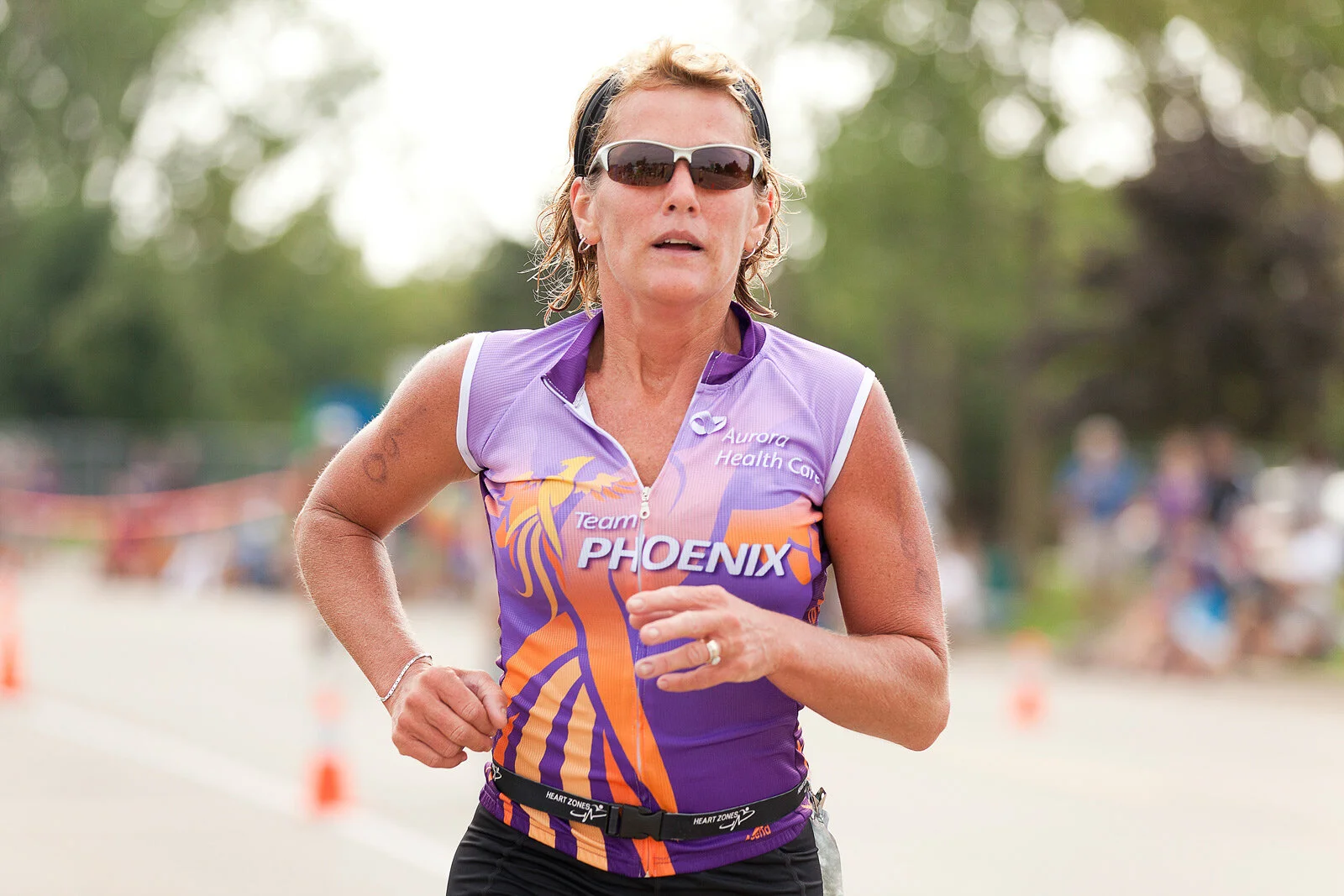A woman running outdoors during a race, wearing sunglasses and a purple and orange sports jersey with 'Team Phoenix' and 'Aurora Health Care' logos.