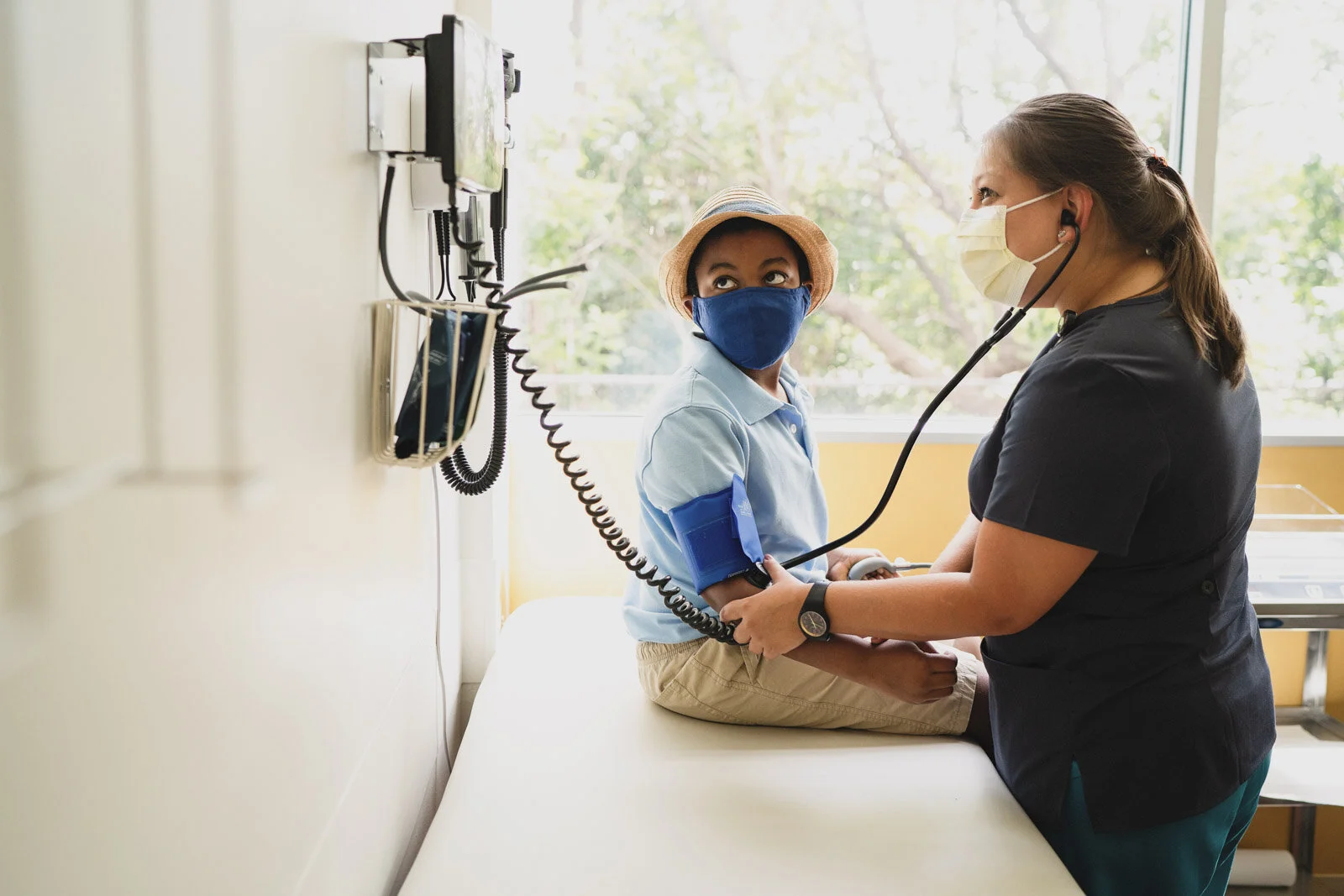 A healthcare professional checks a young boy's heartbeat using a stethoscope in a medical room with large windows and trees outside.