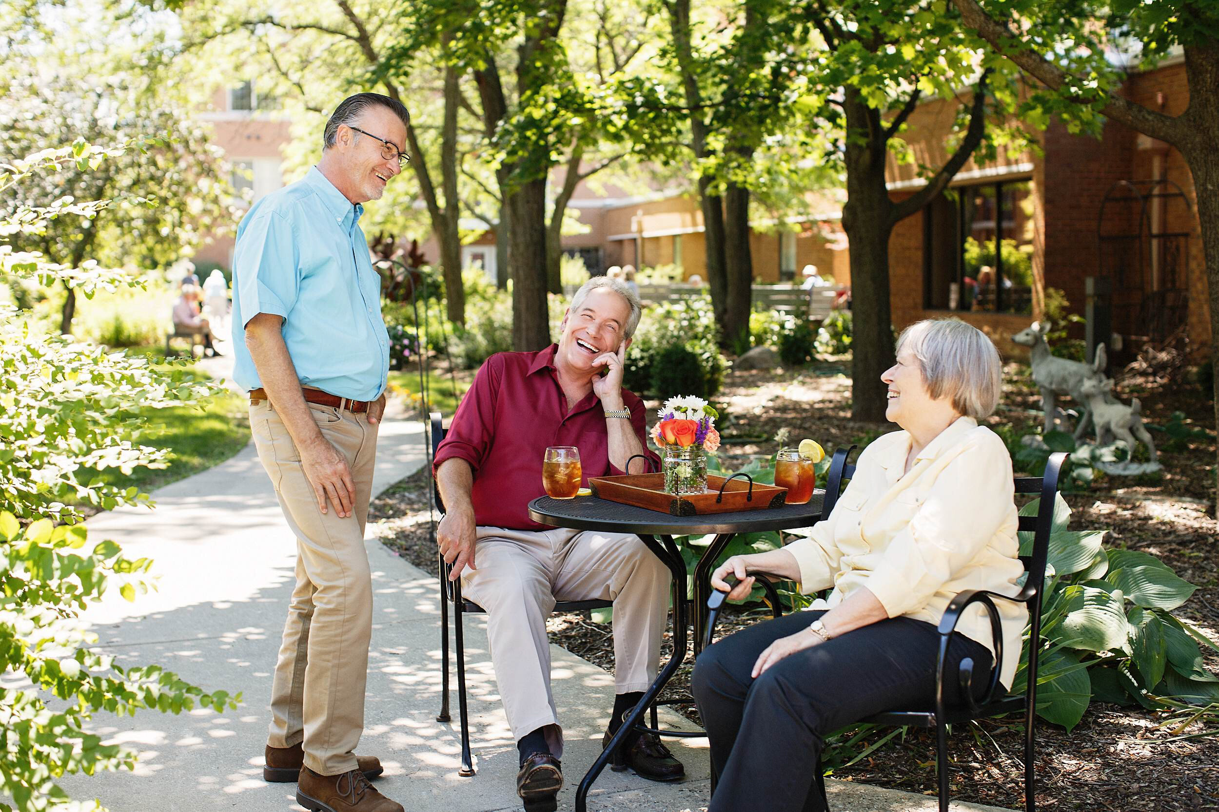 Three elderly people enjoying a sunny outdoor conversation at a park. Two women sitting at a table and a man standing nearby. The women are laughing, one with a cane. The table has a small flower arrangement and drinks. Trees and residential building