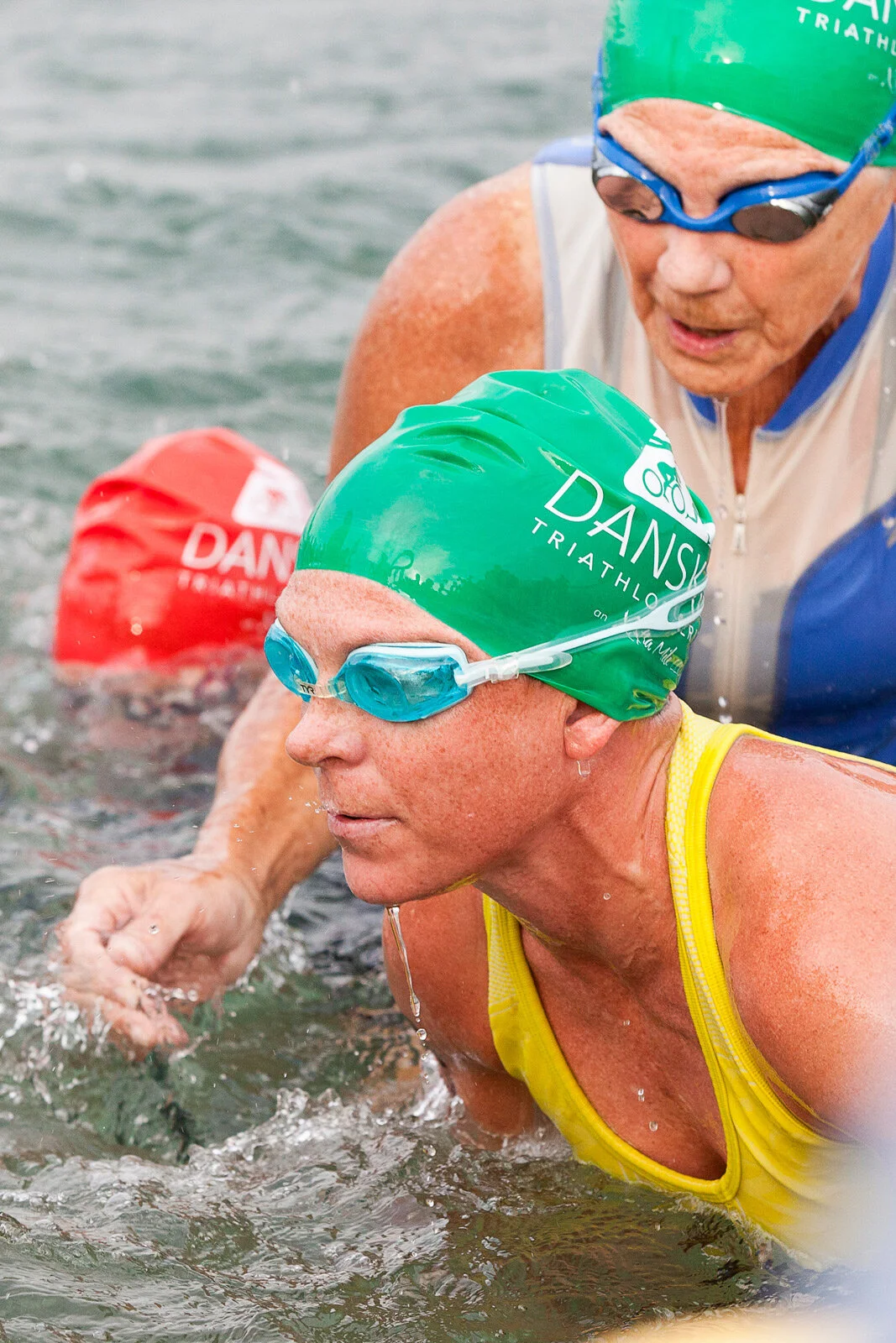 Two women in water, wearing green and red swim caps with 'DANSK TRIATHLON' written on them, and swimming goggles, participating in a triathlon.