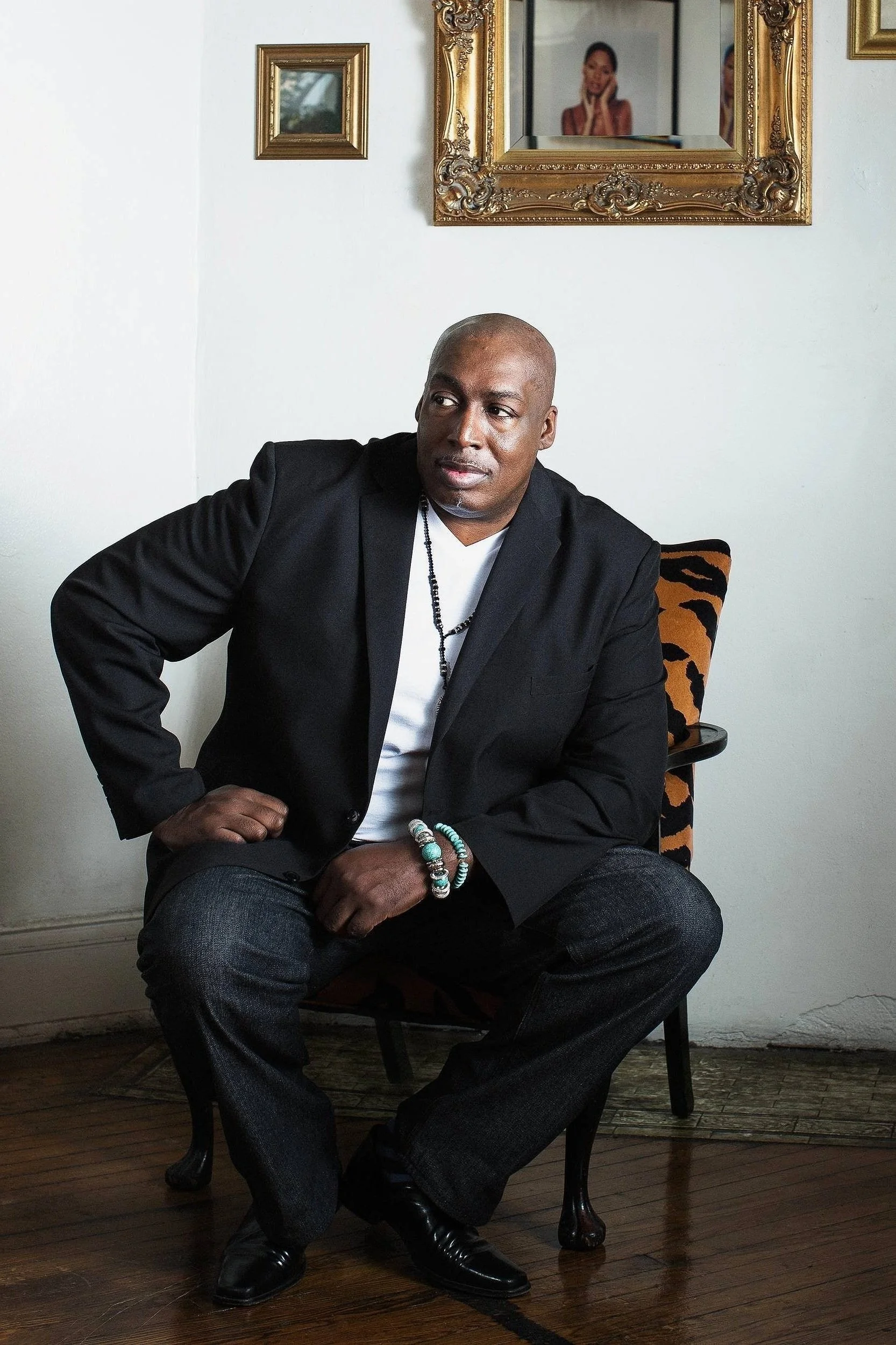 Environmental portrait of a man seated in a chair indoors, wearing a black blazer and white shirt, looking off to the side beneath ornate gold-framed artwork on the wall behind him.