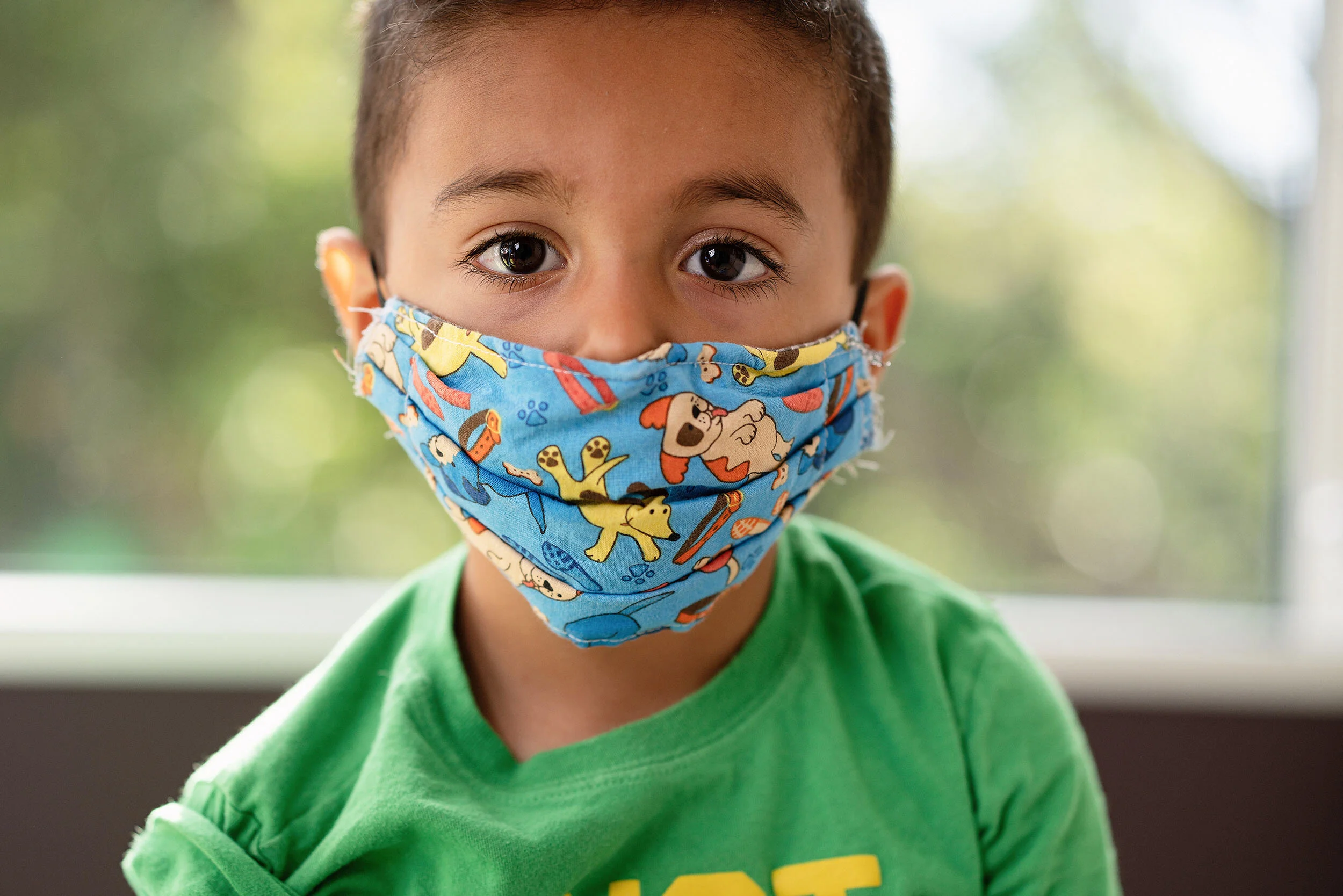 A young boy wearing a colorful cartoon character face mask and a green shirt, standing indoors near a window with blurred greenery outside.