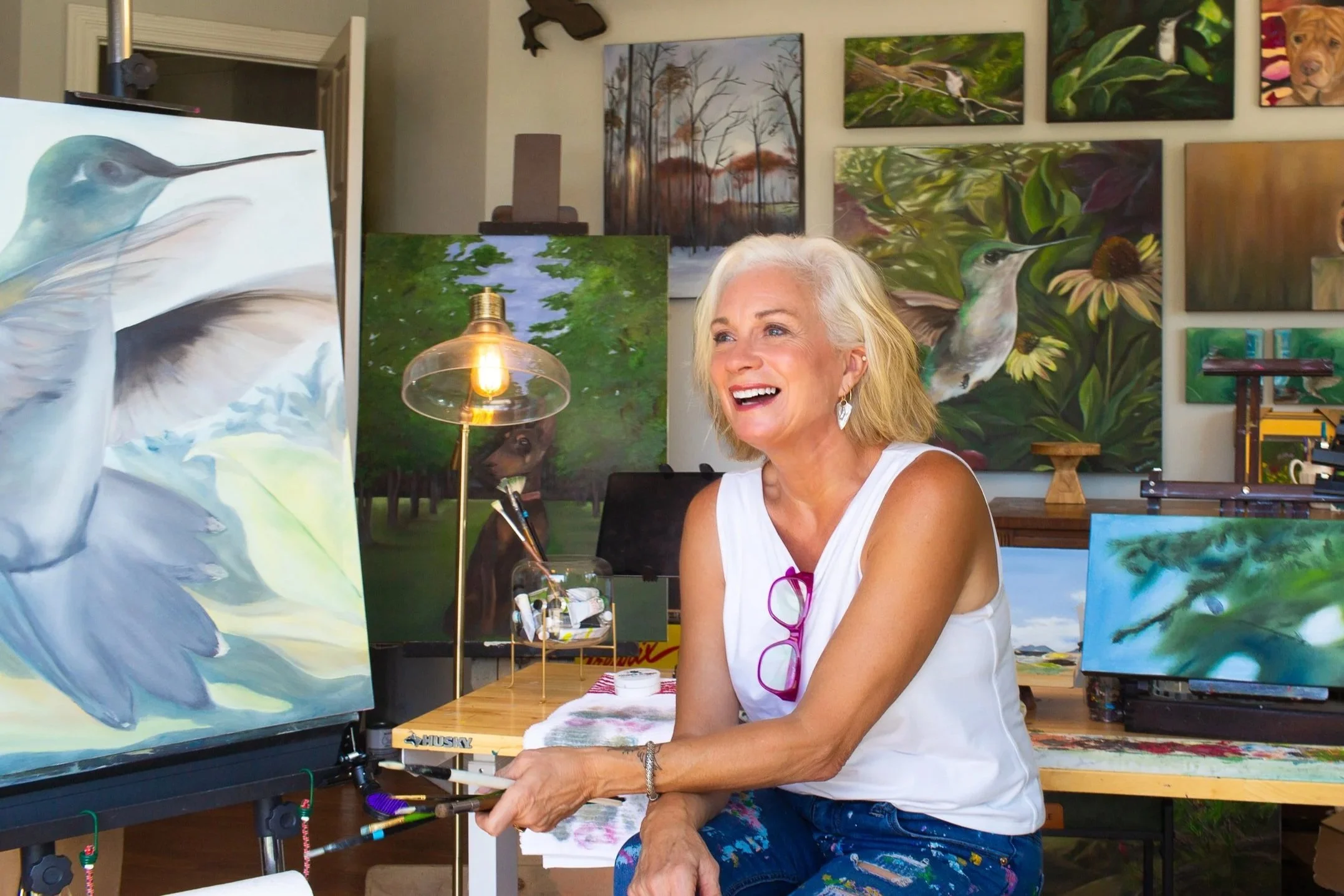 Artist Diana Paxton sitting in her art studio and surrounded by her hummingbird paintings