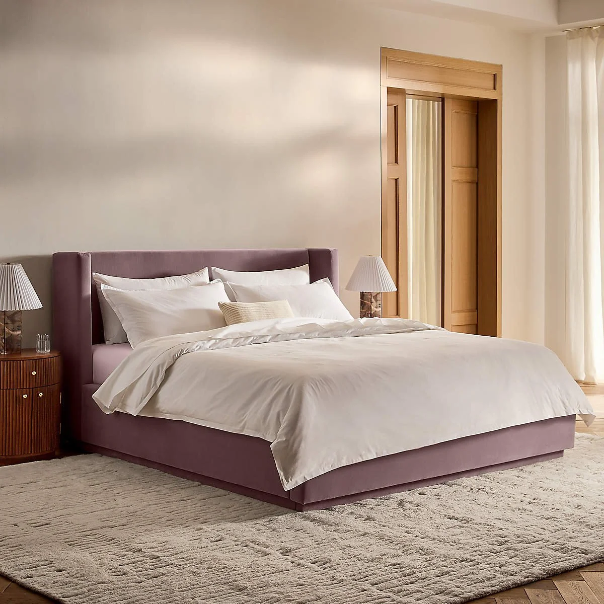 Cama grande con cabecera lila, ropa blanca y una almohada beige, mesitas blancas con lámparas y cortinas claras en habitación moderna y luminosa.