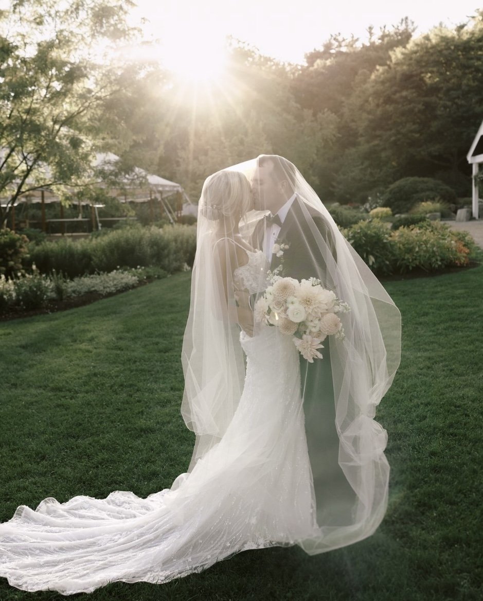 Newlywed couple kissing under a veil in a garden setting during sunset.
