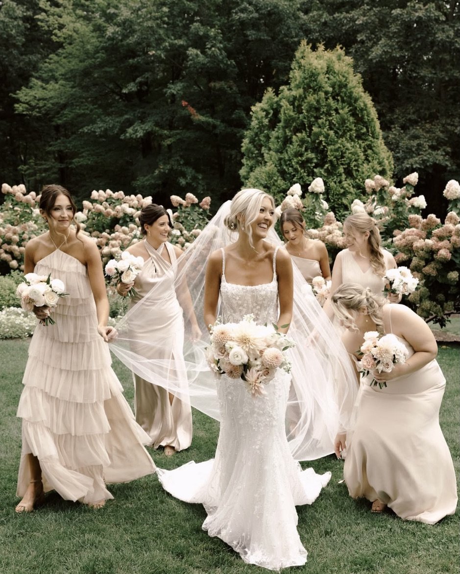A bride in a white dress surrounded by bridesmaids in light-colored gowns, holding bouquets, standing on a grassy area with flowers and trees in the background.