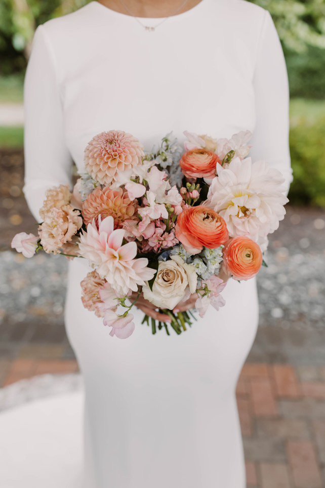 Person in a white dress holding a bouquet of flowers including dahlias, roses, and ranunculus with pastel colors.