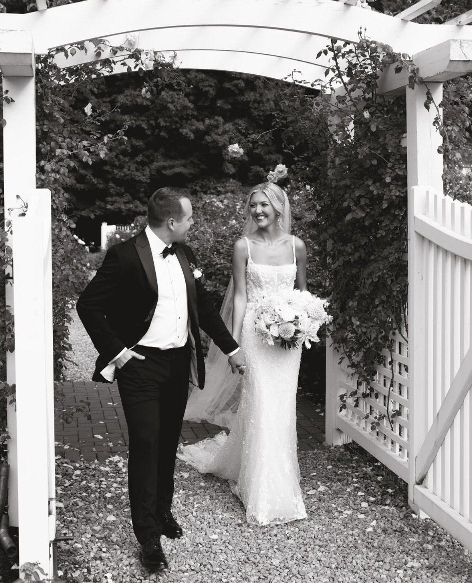 Black and white photo of a couple in wedding attire walking through a garden archway.