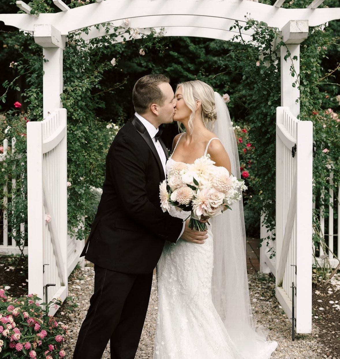 Bride and groom kissing under a white trellis, with greenery and flowers, bride holding a bouquet of white and pink flowers.