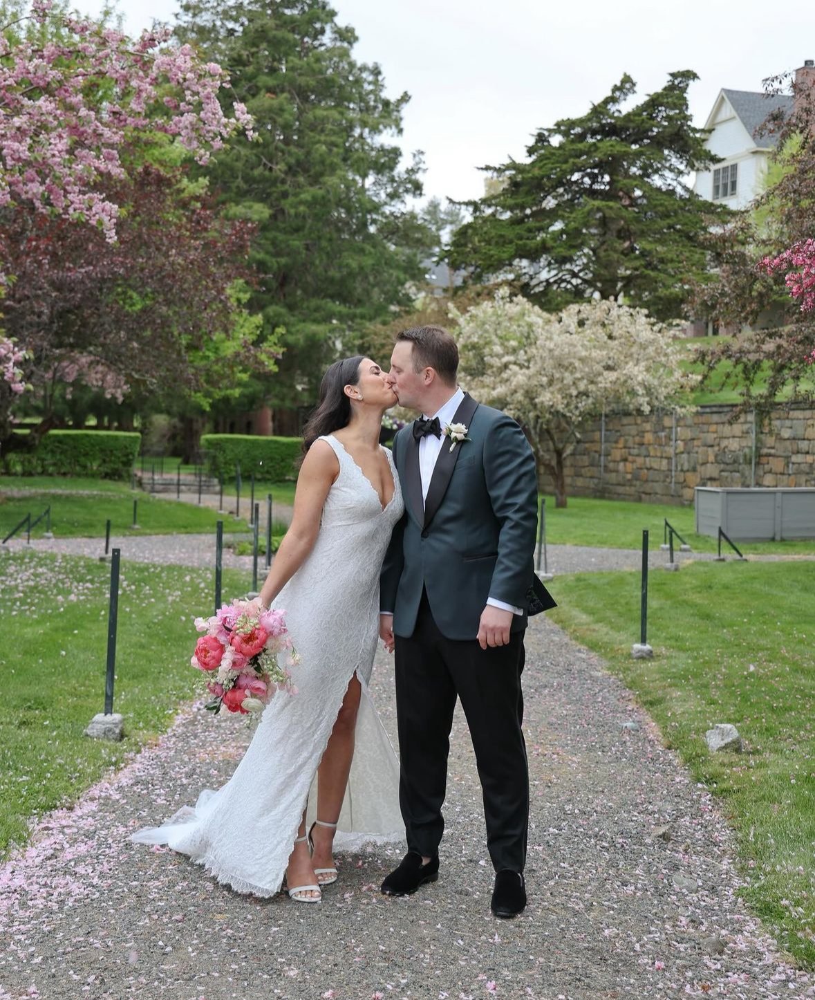 A bride in a white wedding dress and a groom in a dark suit sharing a kiss outdoors, surrounded by green trees and pink flowers, holding a bouquet of pink and white flowers.