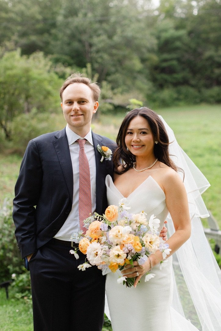 A bride and groom standing outdoors in a garden. The groom is wearing a dark suit with a pink tie and boutonniere, and the bride is in a white wedding dress with a veil, holding a bouquet of yellow, white, and orange flowers.