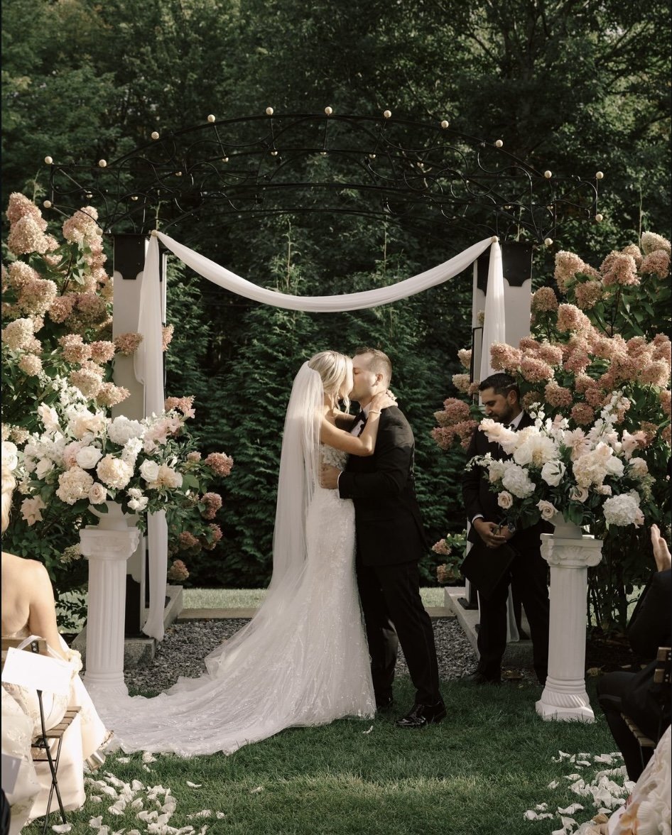 Outdoor wedding ceremony with a bride and groom kissing under an arch decorated with flowers and drapery, surrounded by greenery and floral arrangements.