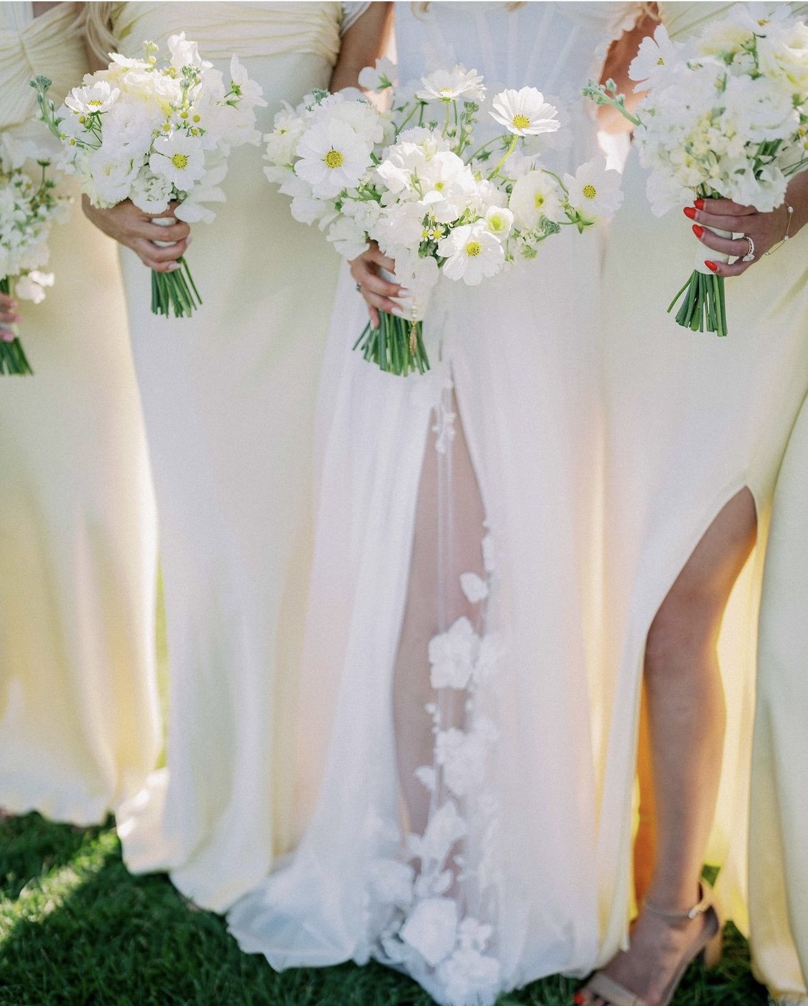 Bridesmaids and bride holding bouquets of white flowers, wearing light-colored dresses, standing on grass.