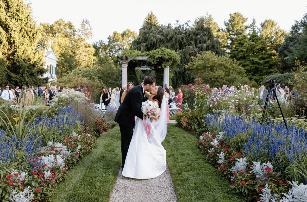 Bride and groom kissing in a garden setting during an outdoor wedding ceremony, surrounded by colorful flowers and guests seated in the background.
