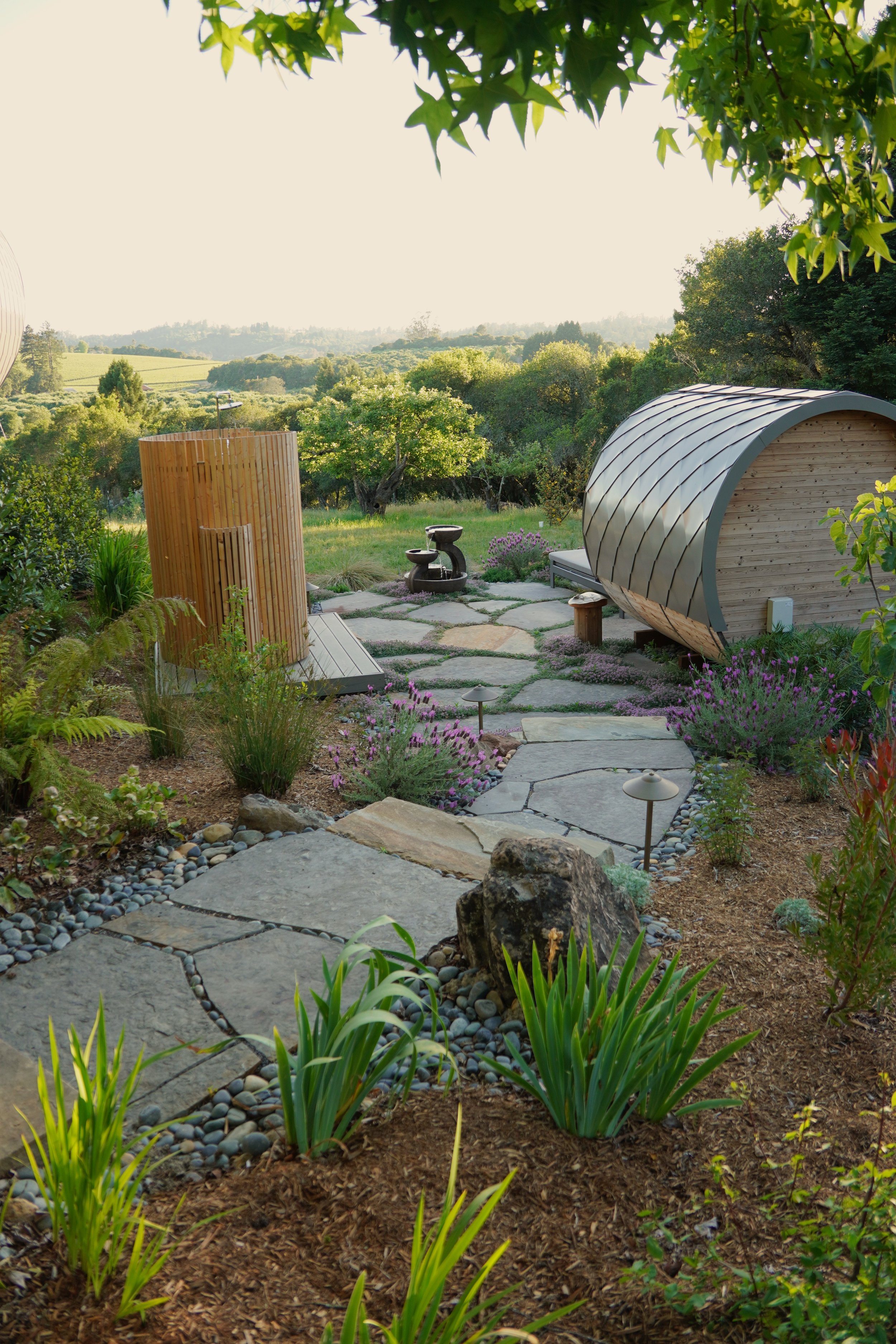 Serene backyard garden with stone pathway, purple flowers, and modern garden structures surrounded by lush greenery and distant hills.
