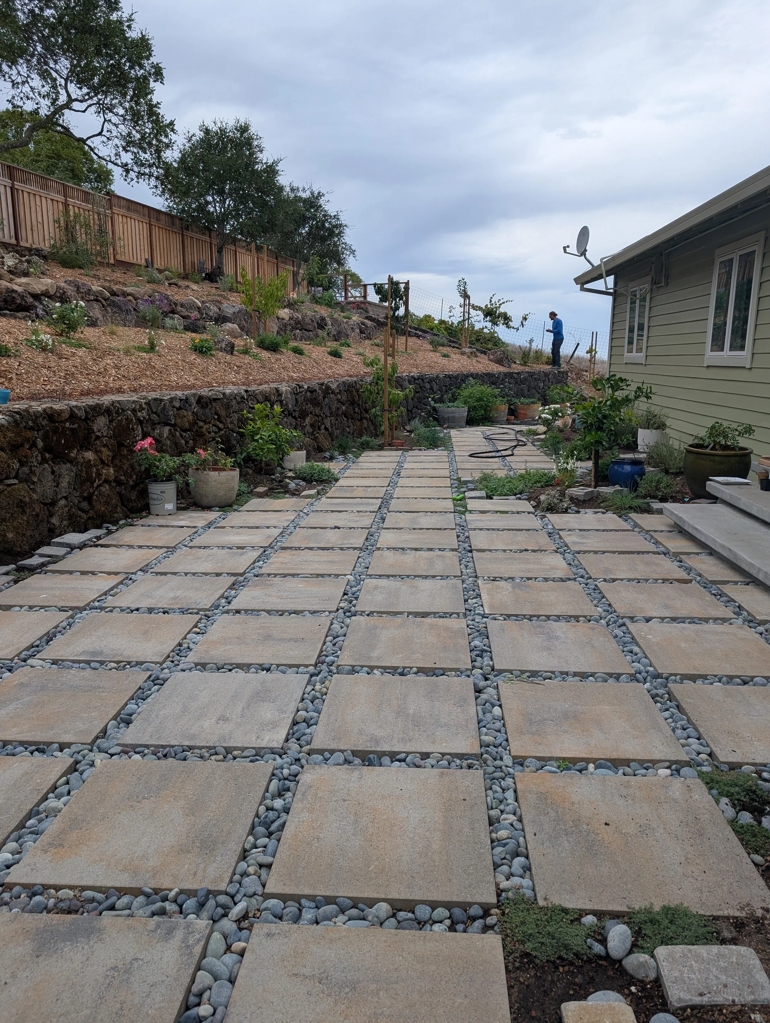 A backyard patio with large square stone tiles arranged in a grid pattern, separated by small gray pebbles. There are potted plants along the edges, a house on the right, and a garden on the hillside in the background. A person is working on the hillside near the fencing.