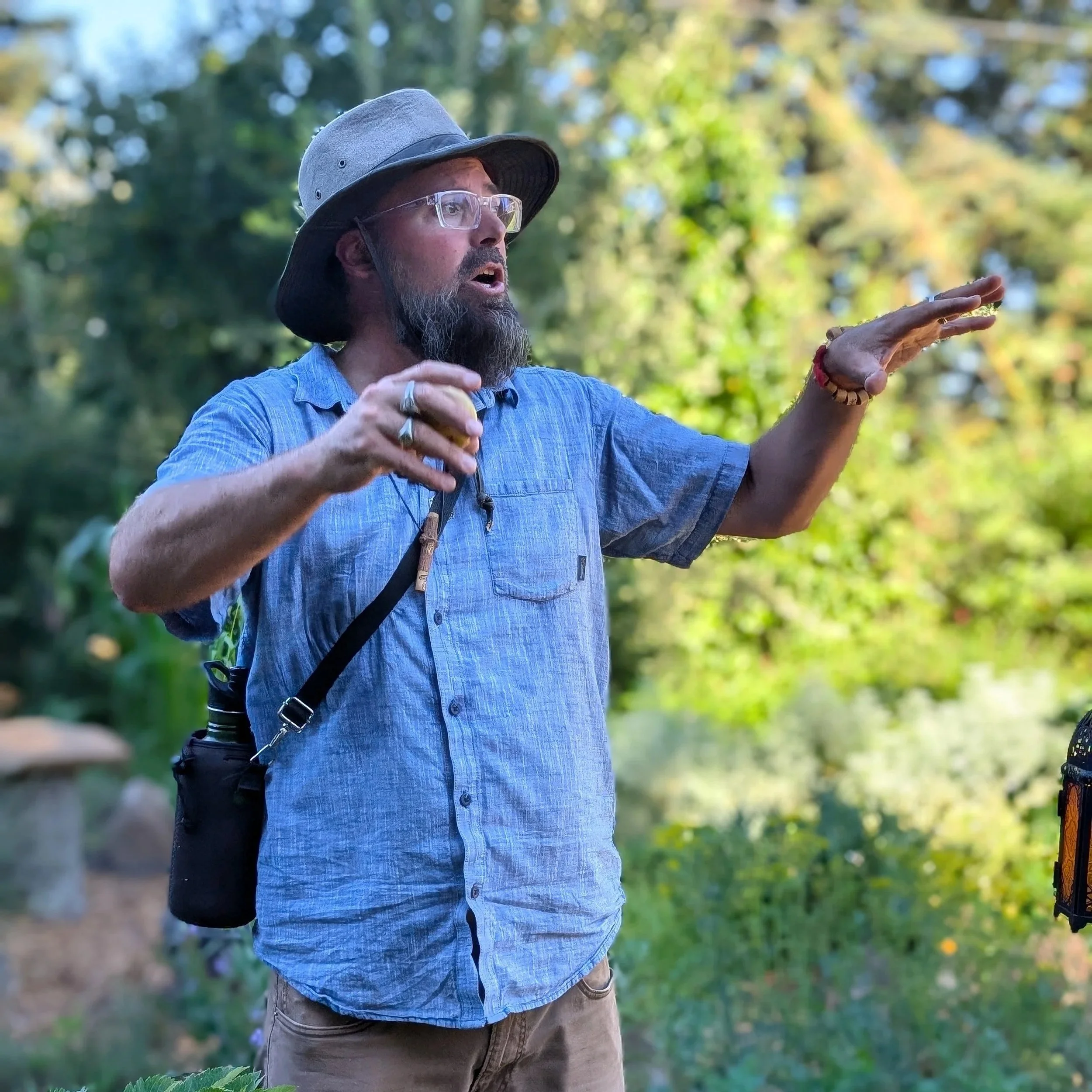 A man with a beard and glasses, wearing a wide-brimmed hat and a blue button-up shirt, explaining or gesturing with his hands in an outdoor, wooded area during daytime.