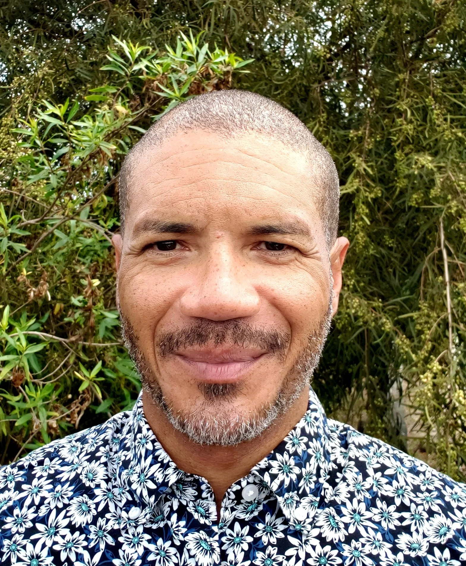 Close-up photo of a smiling man with a bald head, gray beard, and mustache, wearing a floral patterned shirt, standing outdoors with green foliage in the background.