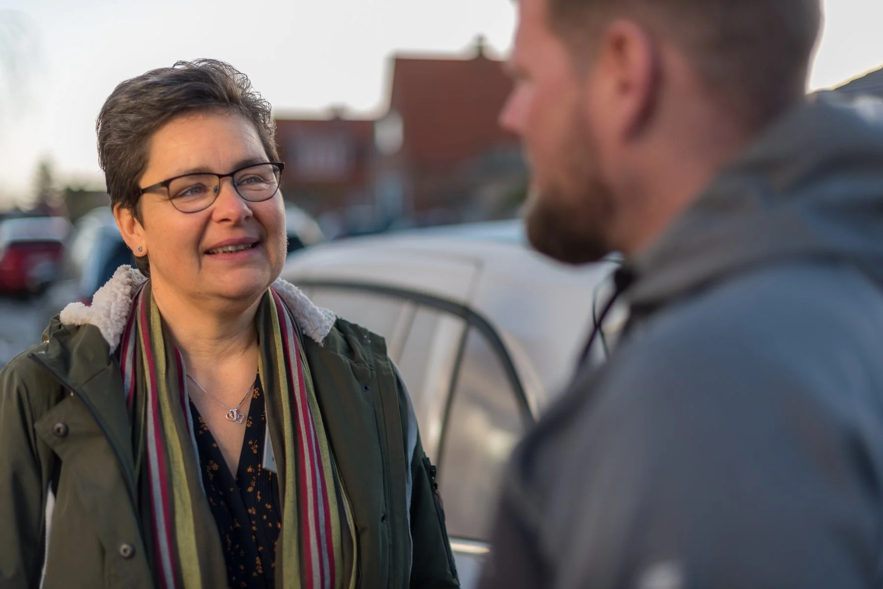 A middle-aged woman with short hair and glasses smiling at a young man with a beard while talking outdoors, with cars and houses in the background.