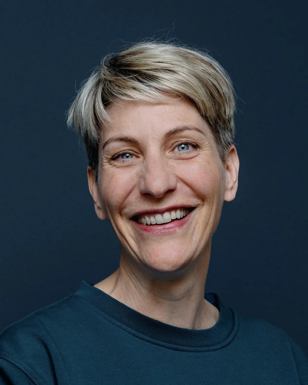 Portrait einer lächelnden Frau mit kurzen blonden Haaren vor einem dunklen Hintergrund