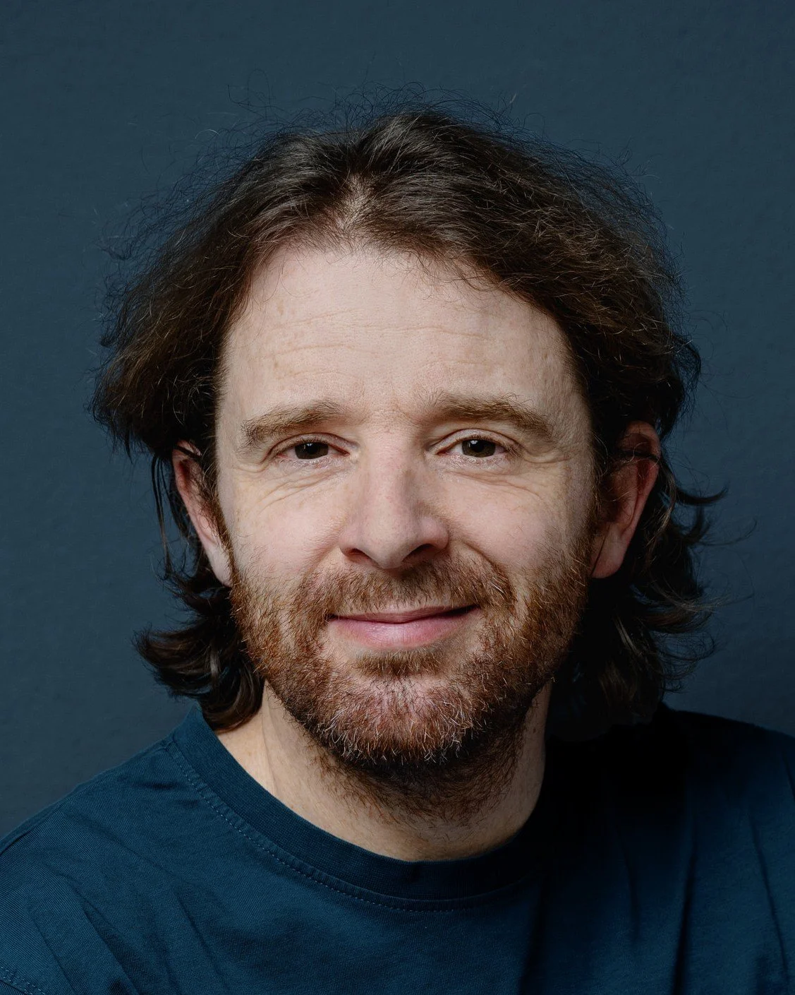 Portrait eines lächelnden Mannes mit langen Haaren und Bart vor einem dunklen Hintergrund