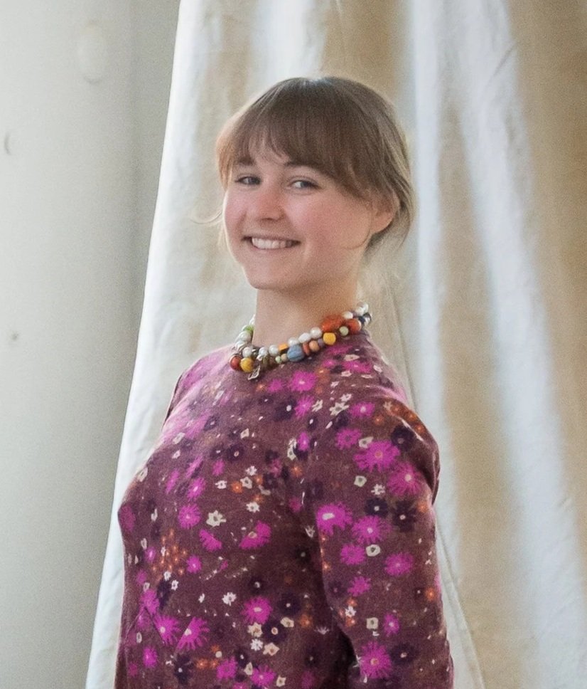 Een jonge vrouw met korte, golvende, lichtbruine haar, lachend, steekt haar arm rustend op een vensterbank, gekleed in een blauwe satijnen blouse met een paisley patroon, een zwart rokje met witte polkadots, een bruine riem met een grote gesp, en draagt meerdere kleurrijke kettingen en sieraden.