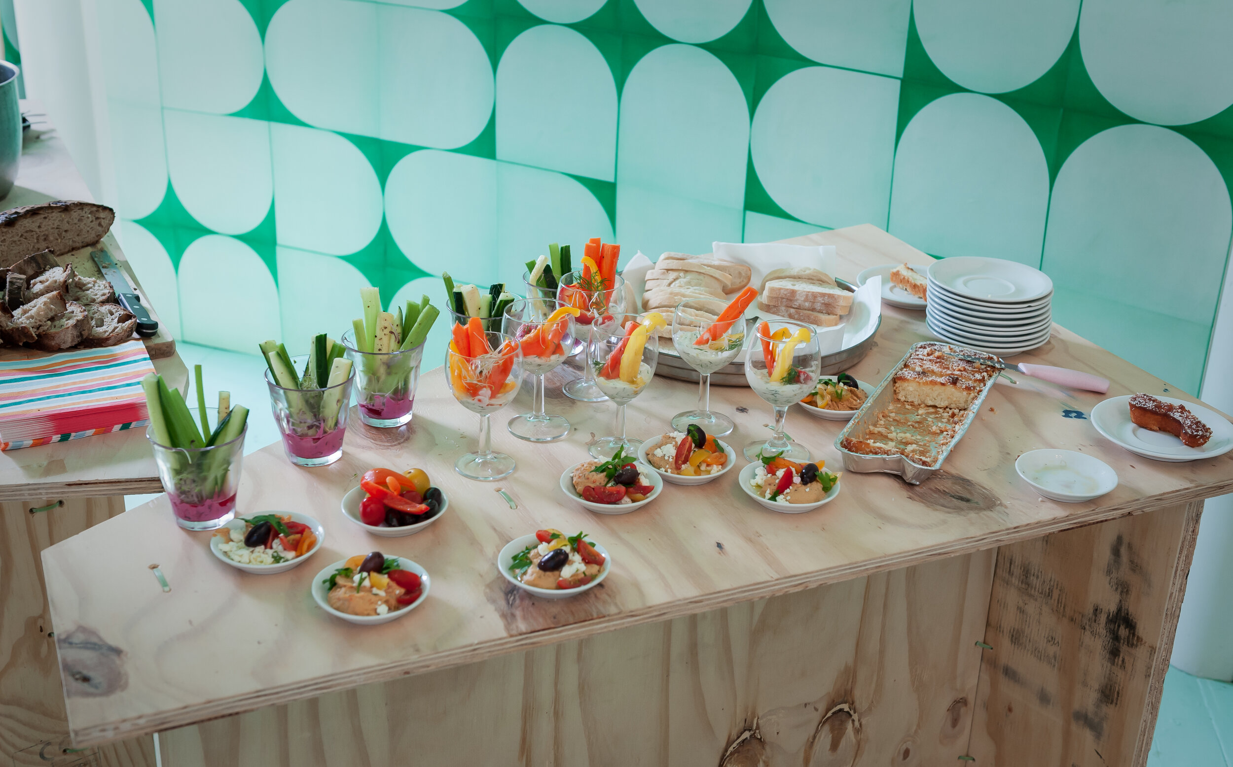 Assorted appetizers on a wooden table, including vegetable sticks in glasses, small plates of salads, sliced bread, a rectangular casserole dish, and stacked plates. Background features green and white geometric wall pattern.