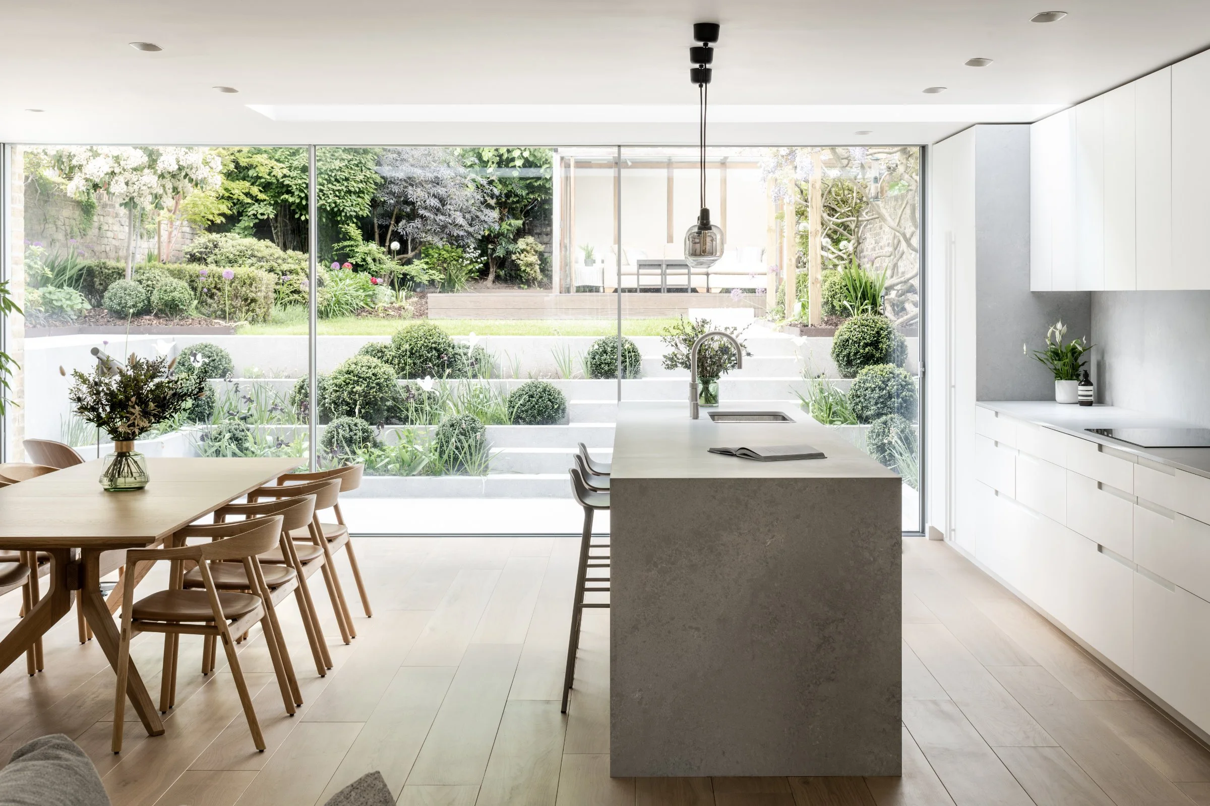 Forgeworks Architects — Modern kitchen with island, dining table, wooden chairs, and large glass windows overlooking a landscaped garden.