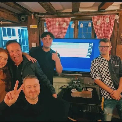 Five people in a cozy room with wooden walls and window with red checkered curtains, standing and sitting around a large screen displaying a list or messages, smiling and making peace signs and thumbs-up gestures.