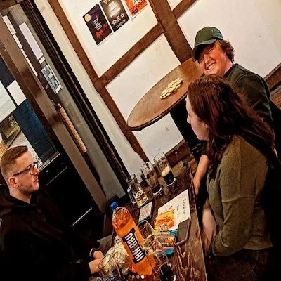 Three people sitting at a table in a casual restaurant or pub, with two men and one woman. They are smiling and engaging in conversation, with drinks and snacks on the table.