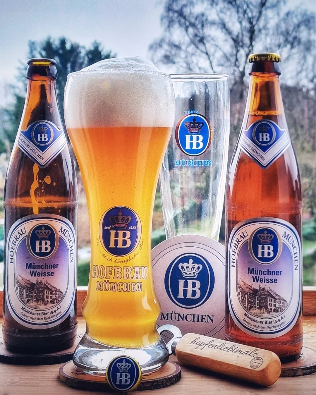 Two bottles of Hofbräu München beer, a filled beer glass, a beer coaster, and a glass with HB logo on a wooden surface with a scenic outdoor background.