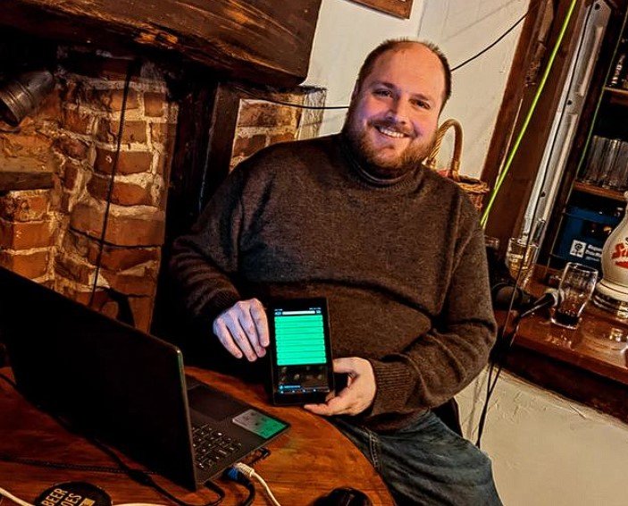 A man with a beard and sweater sitting at a bar holding a smartphone with a glowing screen, smiling at the camera.