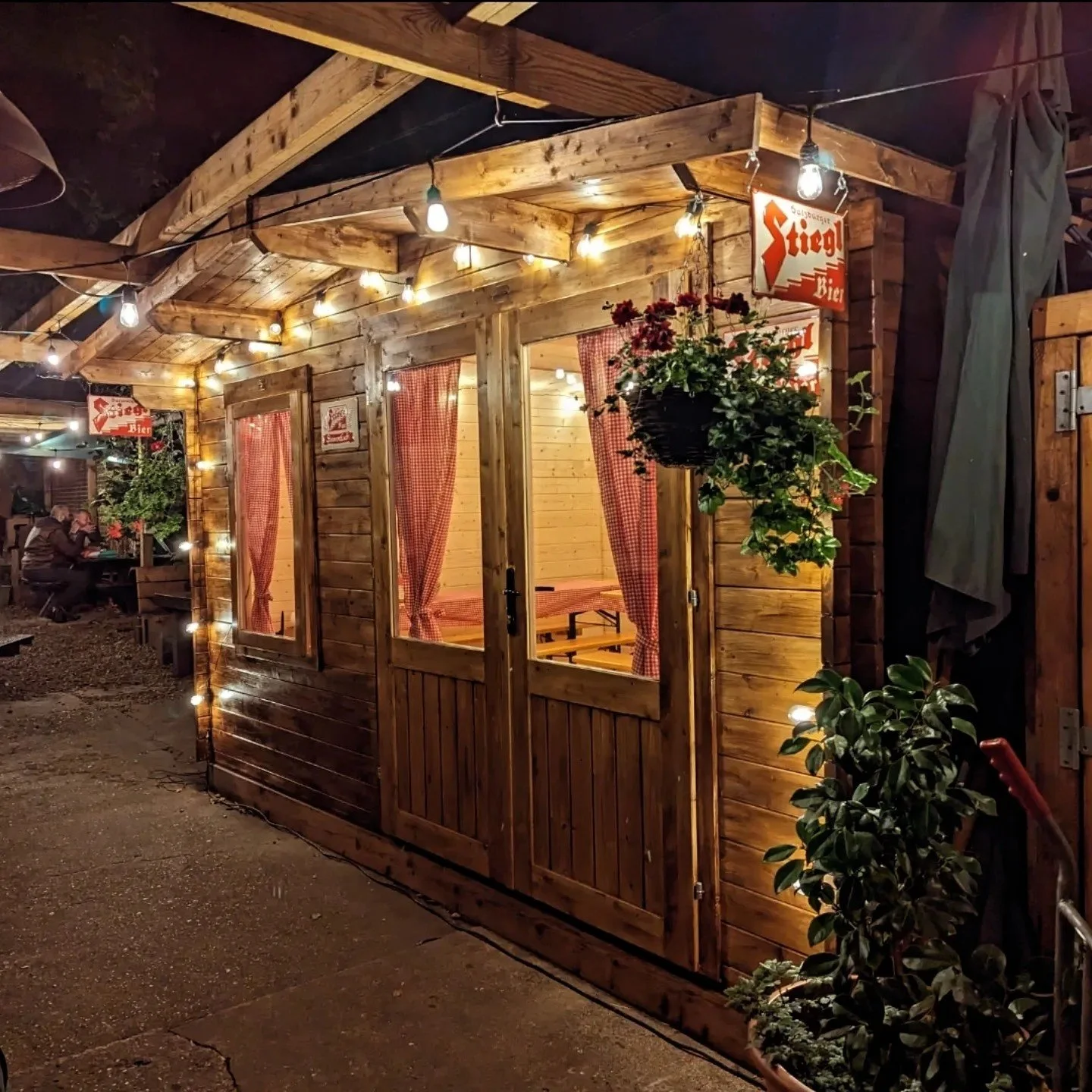 Night view of a cozy wooden cabin with string lights, red checkered curtains, and hanging flower basket, in a lively outdoor setting.