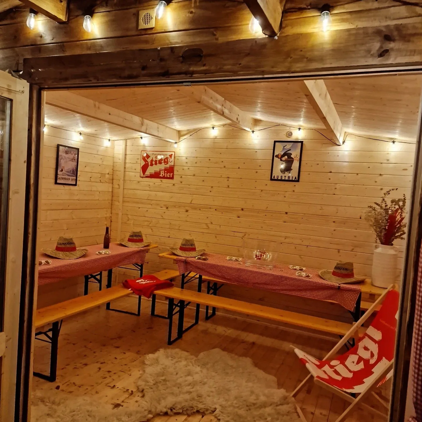 Cozy wooden interior of a small restaurant or café with red checkered tablecloths, straw hats, and vintage posters on the walls, soft string lights, and a white fuzzy rug on the wooden floor.
