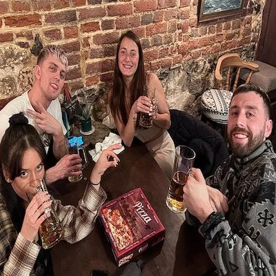 Four friends enjoying drinks around a table in a cozy restaurant or pub with a brick wall background, one woman holding a drink and smiling, a man with a beard smiling, a woman with black hair drinking, and another man with short hair making a peace 