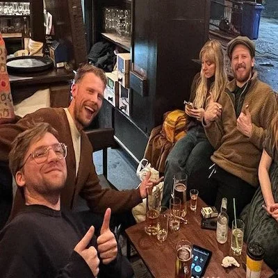 Four people sitting around a wooden table in a bar, smiling and making thumbs-up gestures. Drinks and smartphone on the table, with a girl and a man sitting on a bench in the background.