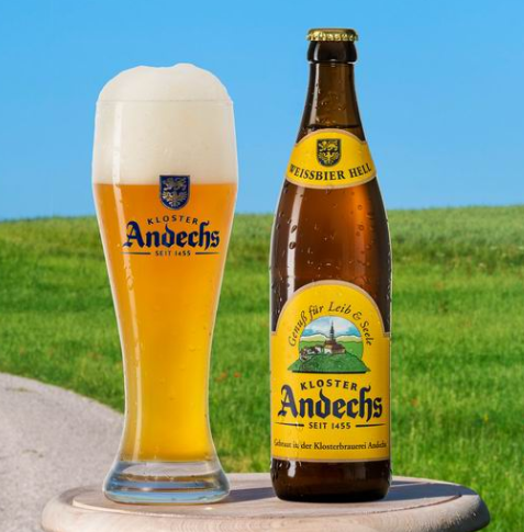 A bottle of Kloster Andechs beer and a tall glass of beer with foam on top, placed on a wooden surface outdoors with a grassy field and blue sky in the background.