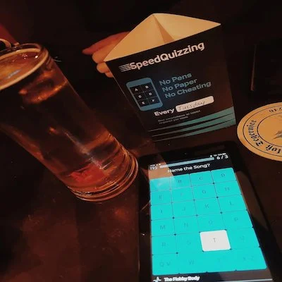 A table at a restaurant or bar with a glass of beer, a smartphone displaying a quiz game, and promotional paper promoting no pens, no paper, and no cheating for a speed quizzing game.